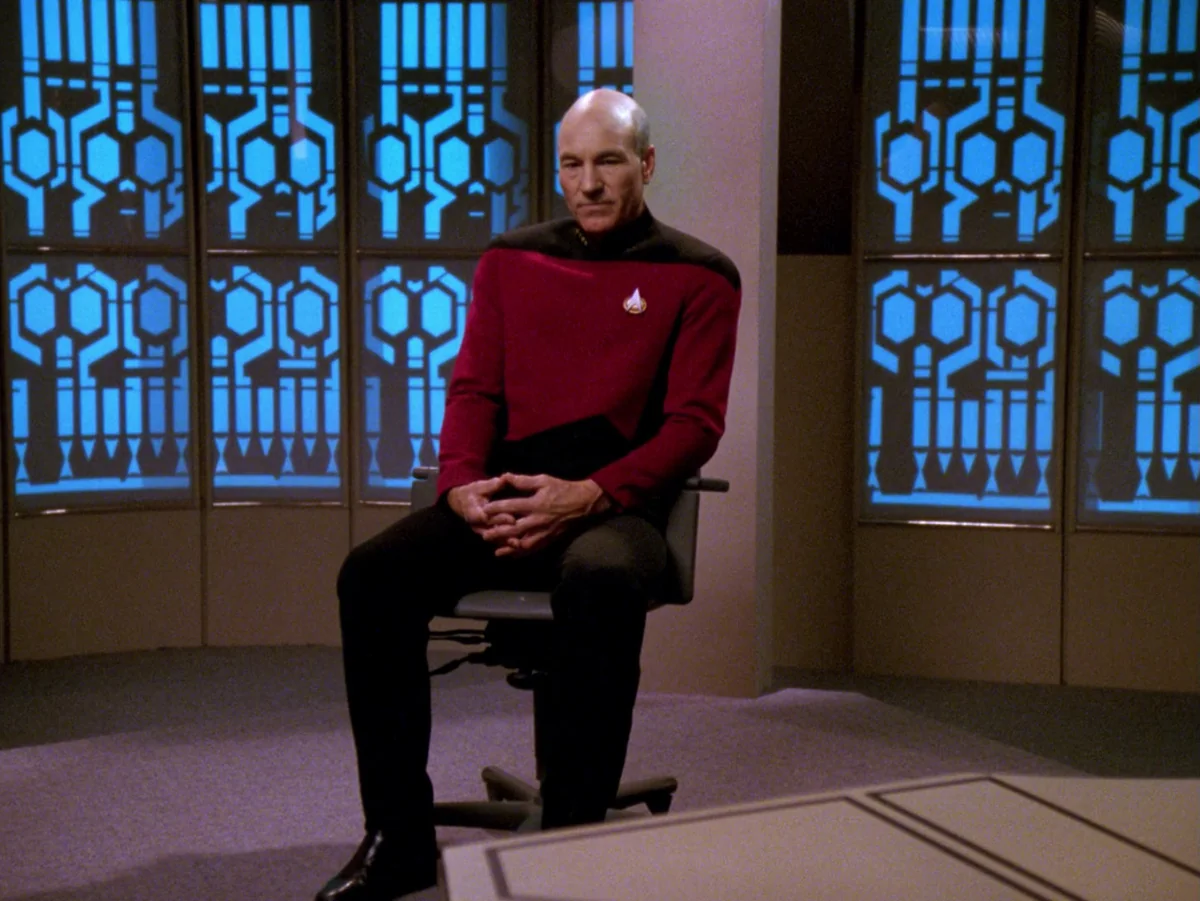 Picard sitting alone on a chair in the hearing room, tall blue-lit windows behind him