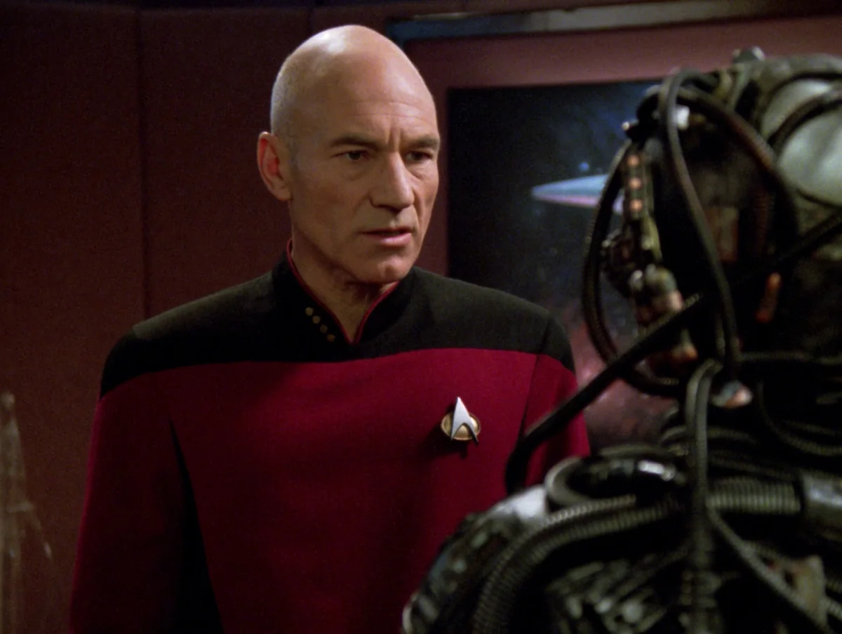 Captain Picard in his red uniform standing face to face with Hugh, a young Borg drone with a mechanical eyepiece