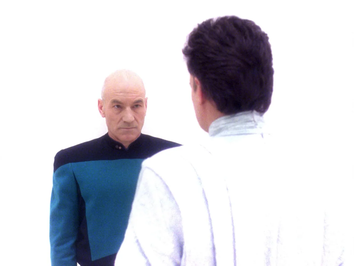 Picard, in the blue-shouldered uniform of a junior science officer, facing Q, who is dressed in white, in a bright white void