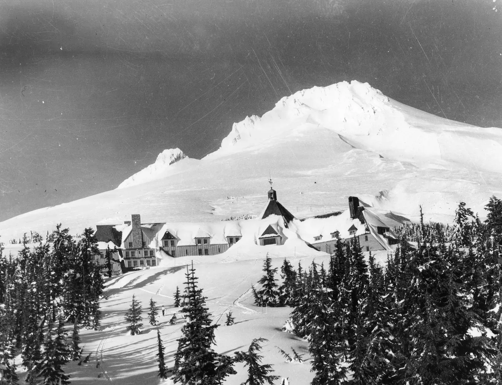 Black-and-white photograph of Timberline Lodge half buried in snow below the peak of Mount Hood, with snow-covered fir trees in front.