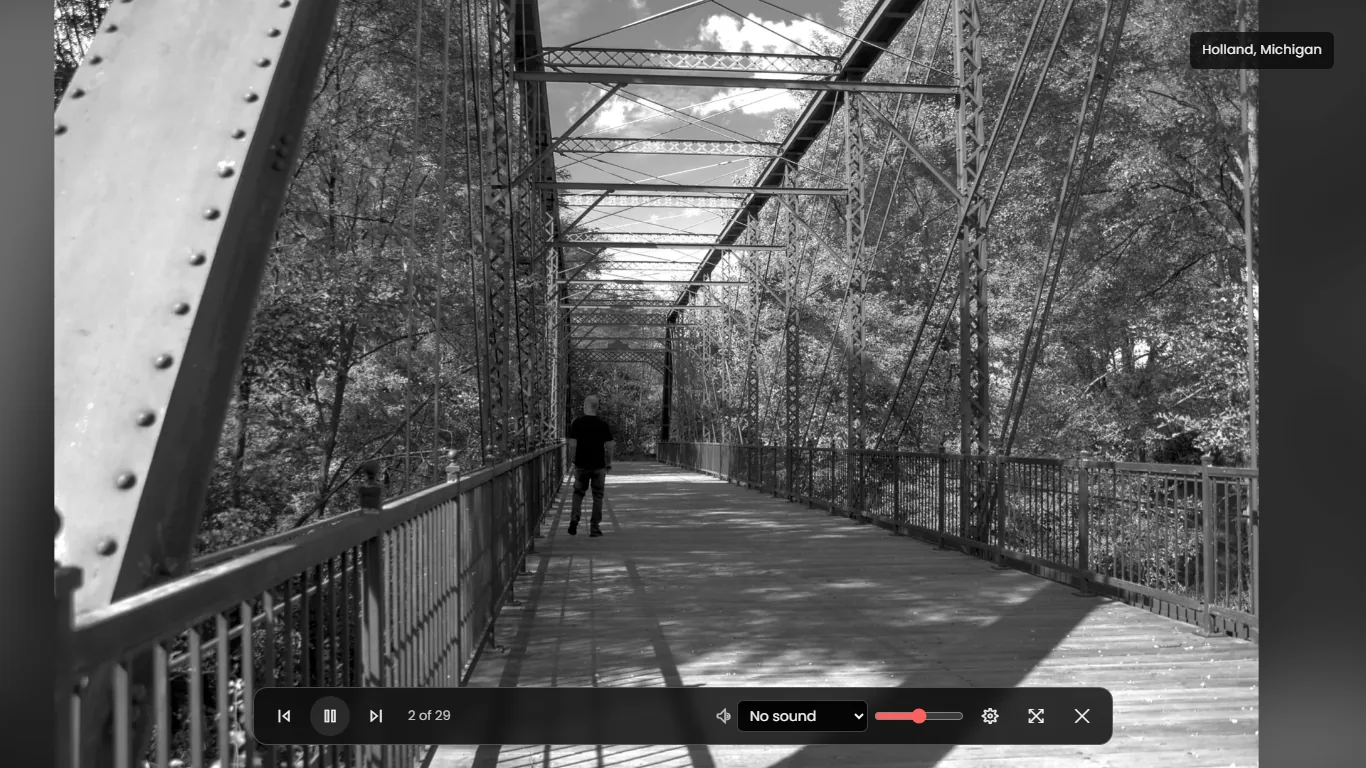 A black and white photograph of a person walking across an iron truss footbridge fills the screen, with a caption reading Holland, Michigan in the top right and a bar of playback, sound and settings controls along the bottom