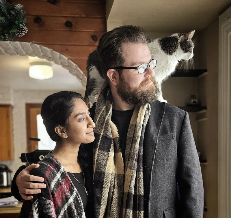 Ken and Neeharika by a window in plaid scarves, his arm around her, a grey and white cat standing on his shoulder