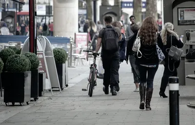 Pedestrians walking away from the camera along a busy city pavement past cafe tables and a parked bicycle