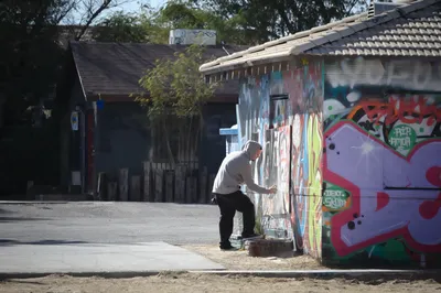 A man in a white shirt bending over to paint on a graffiti-covered single-storey building on a sunny street