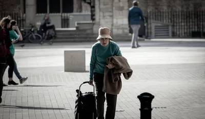 A woman in a sun hat crossing a bright square, a coat over one arm and a wheeled shopping bag in the other hand