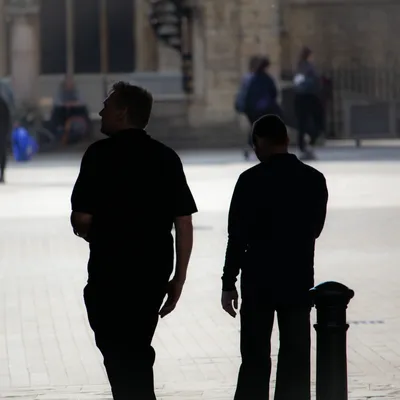 Two men in silhouette crossing a sunlit square, backlit against pale paving