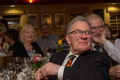 An older man in glasses and a tie at a busy restaurant dinner table, mid-conversation, wine glasses and flowers in front of him