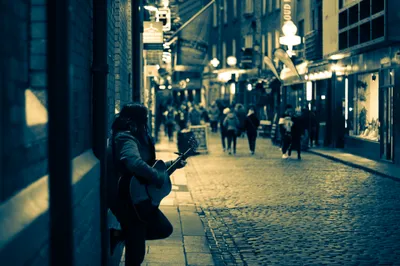 A busker playing guitar against a shopfront on a cobbled street at dusk, the lit lane running away behind them