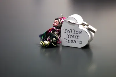 A small white heart-shaped tag reading Follow Your Dreams beside a tiny knitted doll, on a dark grey surface