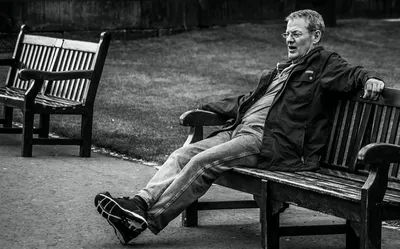A man stretched out along a park bench in the afternoon, arms spread across the backrest, black and white