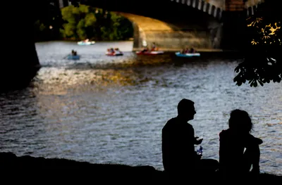 Two people sitting in silhouette on a riverbank under a stone bridge, kayaks on the water beyond