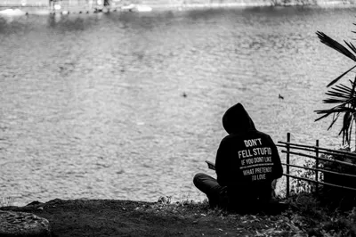 A person in a hooded top sitting alone on a riverbank looking out over the water, a slogan printed across the back of the hood