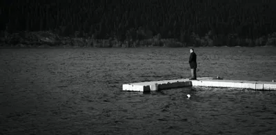 A lone figure standing at the end of a floating jetty on dark water, wooded hills behind, black and white