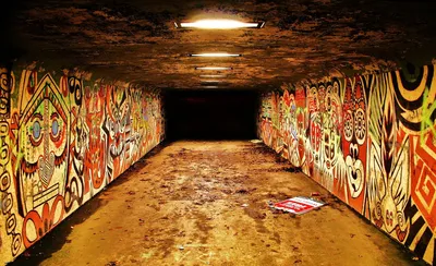 A pedestrian underpass painted on both walls with large faces and patterns in red, yellow and black, lit by ceiling lamps
