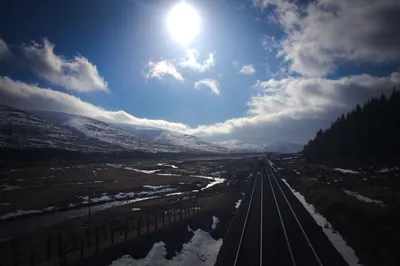 Railway tracks running toward snow-covered Scottish hills under a bright sun and scattered clouds