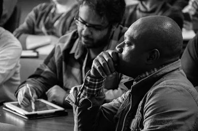 Black-and-white photograph of adult students in a classroom, one deep in thought and another taking notes on a tablet