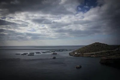 A moody grey sea under heavy clouds, distant fish-farm pens near a rocky headland