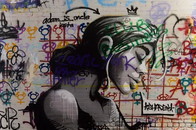 A painted mural of a woman's face in black, white and grey, eyes closed, on a brick wall covered in coloured tags and gender symbols, with a sprayed handle and arrow beside her