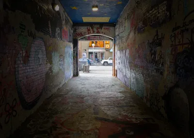 A dim graffiti-covered passage with a painted starry ceiling, opening onto a sunny street with a parked car