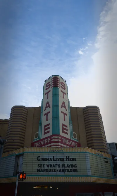 The vertical Art Deco sign of the State Theatre in Ann Arbor rising above its marquee, against a pale blue sky
