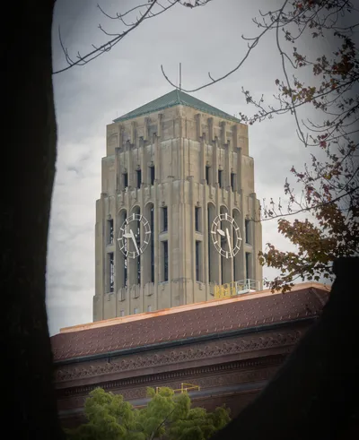 Burton Memorial Tower at the University of Michigan, seen past a red-brick roof and autumn leaves