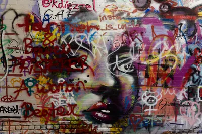 A painted woman's face with red lips almost buried under layers of coloured tags and names on a brick wall
