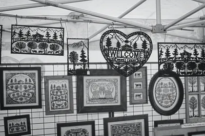 A market stall wall hung with framed black papercut pictures of trees, houses and a heart reading Welcome, black and white